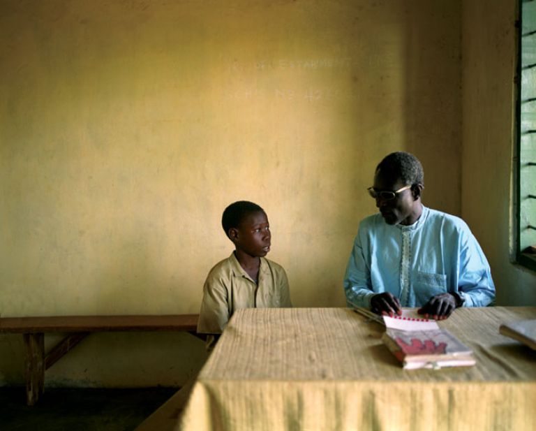 Image 11Orphaned student with blind teacher.Dico Abulta is 15 years of age. At the time of his birth, himself and his mother lived on the Ivory Coast. During their time there, she contracted HIV. Soon after that, she returned to Damongo without her son as she knew her inevitable fate. It was not until Abulta was 7 years of age that a distant relative came in search of him. Abulta now lives with his elderly grandmother who he has to provide for by working as a farm labourer after school. Copyright Paul V. Kelly 2003